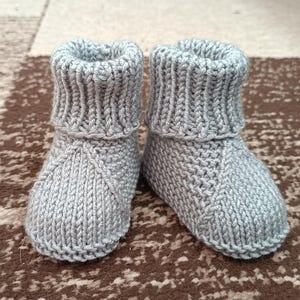 May include: A pair of gray knitted baby booties. The booties have a ribbed cuff and a textured knit pattern. The booties are designed to keep a baby's feet warm and cozy. The booties are made of soft yarn.