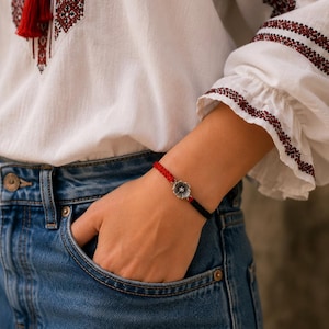 May include: A bracelet featuring a braided red and black cord with a central silver-toned metal flower charm. The charm has a detailed petal design with a small circular center. The cord is adjustable and sits securely around the wrist area.