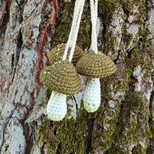 May include: Three crocheted mushroom ornaments with green and white accents hanging from a tree trunk. The mushrooms have a brown cap and a white stem.