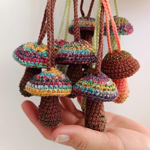 May include: A collection of handmade crochet mushroom pendants in various colors. The mushroom caps feature vibrant rainbow hues, while the stems are brown. Each pendant is strung on a brown or green cord, with green beads around the stem.