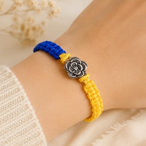 May include: A braided cord bracelet featuring a vibrant blue section and a bright yellow section, connected by a central silver-toned metal charm shaped like a detailed flower with layered petals and an oxidized finish.