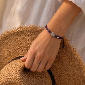 May include: A purple braided cord bracelet featuring a central silver-toned sea turtle charm and two small silver-toned spherical beads. The bracelet is secured with a sliding knot closure and is worn on a wrist.