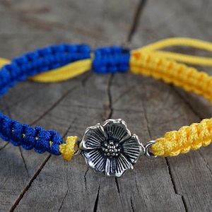 May include: A macrame bracelet featuring blue and yellow braided cord, with a silver-colored flower charm at the center. The bracelet's adjustable design allows for a comfortable fit. The color scheme evokes a sense of vibrancy and style.