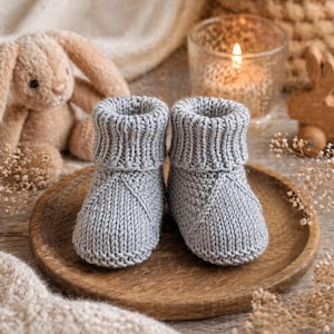 May include: A pair of gray knitted baby booties with folded cuffs, displayed on a wooden plate. A plush bunny and a lit candle are in the background, creating a cozy, handmade theme.