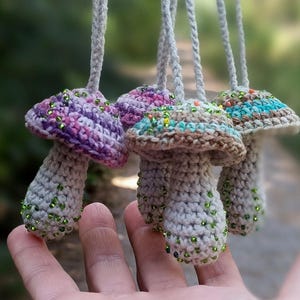May include: A collection of four small, handmade crochet mushroom ornaments hanging from braided yarn loops. Each mushroom features a light-colored stem and a cap decorated with variegated yarn colors and small, scattered green glass beads.