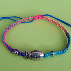 May include: A colorful macrame bracelet featuring a silver fish charm. The bracelet is made with blue, pink, green, and purple threads. Adjustable blue cords allow for a custom fit. A fun, nautical-themed accessory.