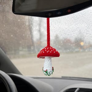May include: A crocheted mushroom car ornament hanging from a rearview mirror. The mushroom has a red cap with white stitched spots, a white stem, and a red braided hanging cord. The base of the stem is decorated with small green and red beads.
