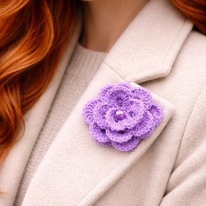 May include: A handmade, lavender crochet flower brooch with a sparkling center. The floral accessory is pinned to a light beige coat. The flower's petals are intricately designed, adding a touch of elegance to the garment.