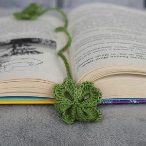 May include: A green, crocheted four-leaf clover bookmark rests in an open book. The bookmark's stem extends upwards, with another clover at the top. The book's pages are cream-colored, and the cover is visible with a blue and purple design.