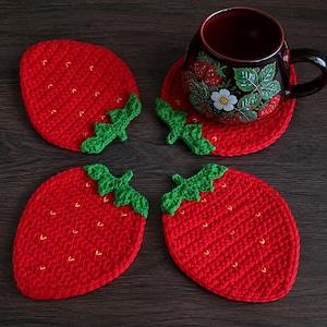 May include: A set of four red crochet coasters shaped like strawberries with green leaves and small yellow embroidered seeds. One coaster supports a dark ceramic mug decorated with a strawberry plant pattern featuring red berries and white flowers.