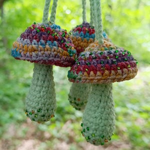 May include: Three handmade crochet mushroom ornaments. The mushroom caps are multicolored with shades of purple, blue, yellow, and red, and are adorned with small green and red beads. The stems are light green, and the ornaments hang from green cords.