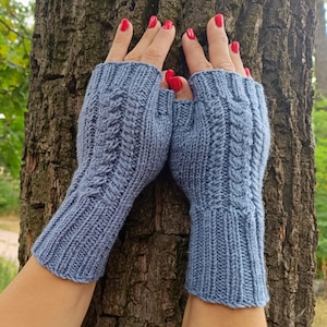 May include: A pair of blue fingerless gloves with a cable knit design. The gloves are made of a soft, knitted material and are designed to keep hands warm while leaving fingers free. The gloves are a muted blue color.