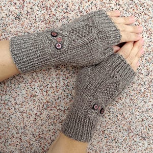 May include: A pair of grey fingerless gloves knitted with a ribbed cuff and a textured cable pattern. Each glove features two small, dark circular buttons with pink centers attached to the outer surface, providing a decorative detail to the knit.