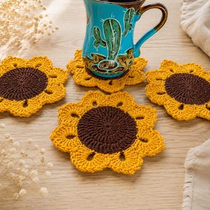 May include: Three round crochet coasters shaped like sunflowers with dark brown centers and bright yellow petals. One coaster supports a turquoise ceramic mug featuring a green cactus illustration. The coasters are arranged on a light wood surface.