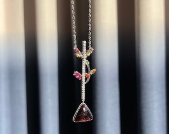 May include: A silver chain necklace with a pendant featuring a triangular red gemstone and a delicate branch design with small colourful gemstones.
