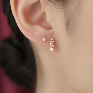 May include: A close-up of a person's ear adorned with two earrings. One is a small, round stud with a central diamond surrounded by smaller stones. The other is a gold drop earring with a pearl and multiple small diamonds.