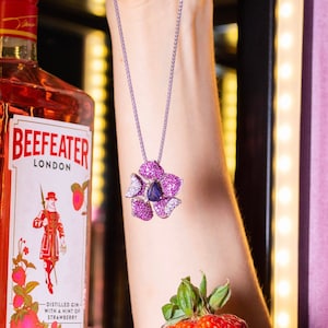May include: A bottle of pink strawberry flavored Beefeater London gin with a silver chain necklace featuring a purple flower pendant with a blue gemstone. A red strawberry is being held in a hand.