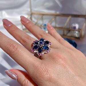 以下が含まれることがあります： 花の形をした、大きな青い宝石と小さなピンクの宝石が囲まれた銀の指輪です。