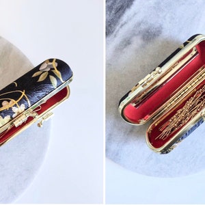 Könnte beinhalten: Zwei schwarze und goldfarbene Haarnadel-Etuis mit roter Innenausstattung. Ein Etui ist geschlossen, das andere ist offen und zeigt goldene Haarnadeln. Die Etuis haben ein Blumenmuster und goldfarbene Beschläge. Die Etuis befinden sich auf einer weißen Oberfläche.