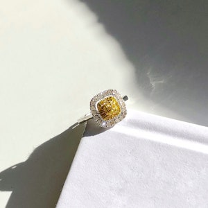 Puede incluir: Un anillo de forma cuadrada con una piedra central de diamante amarillo y blanco rodeada por un halo de diamantes m&aacute;s peque&ntilde;os.