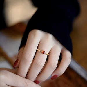 May include: A gold-colored ring with a heart-shaped, reddish-orange gemstone at the center. The ring has delicate floral or leaf-like details on the band. The ring is worn on a finger, with the hand and part of a black sleeve visible.