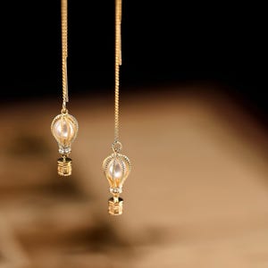 May include: A pair of gold-tone drop earrings with a pearl-like hot air balloon charm. The earrings are hanging from a gold chain.