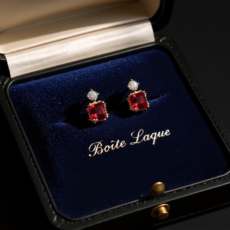 May include: A pair of gold-toned earrings with square, red gemstones and small, round, clear stones. The earrings are displayed in a black jewelry box with a blue velvet interior. The text "Bo&icirc;te Laque" is written in gold script.