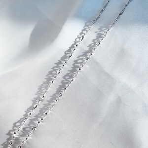 May include: A delicate silver necklace with an elongated oval link chain. The necklace is displayed on a white, textured fabric, with shadows adding depth. The chain is approximately 18 inches long.