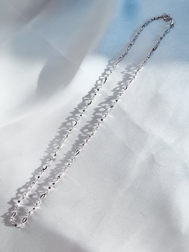 May include: A delicate silver necklace with an elongated oval link chain. The necklace is displayed on a white, textured fabric, with shadows adding depth. The chain is approximately 18 inches long.