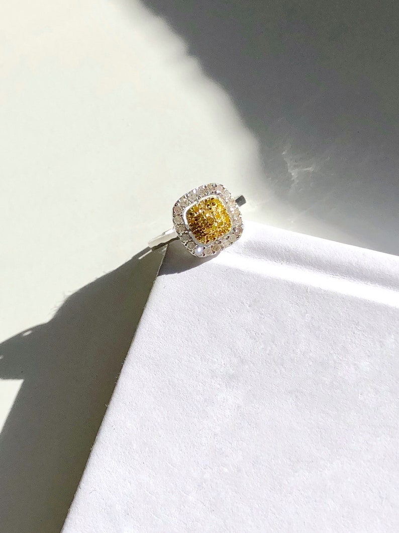 Puede incluir: Un anillo de forma cuadrada con una piedra central de diamante amarillo y blanco rodeada por un halo de diamantes m&aacute;s peque&ntilde;os.