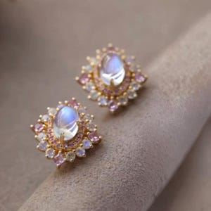 Puede incluir: Un par de pendientes de bot&oacute;n dorados. Cada pendiente presenta una piedra preciosa central, ovalada e iridiscente, rodeada de piedras rosas y blancas. Los pendientes tienen forma cuadrada con un dise&ntilde;o floral.