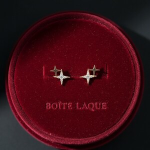 May include: Gold-coloured star-shaped stud earrings with small, sparkling stones, presented in a red velvet jewellery box. The box has the text "BOITE LAQUE" embossed on the front.