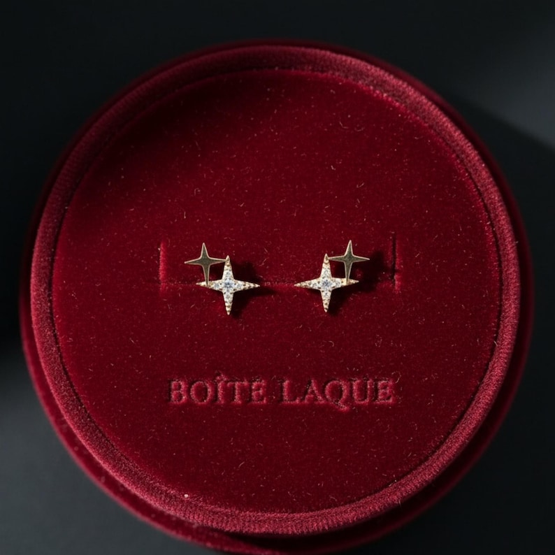 May include: Gold-coloured star-shaped stud earrings with small, sparkling stones, presented in a red velvet jewellery box. The box has the text "BOITE LAQUE" embossed on the front.