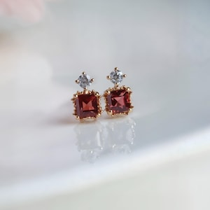 May include: A pair of gold-toned square earrings. Each earring features a large, square, deep red gemstone, topped with a smaller, clear, square-cut stone. The edges of the earrings have a decorative, textured design. The earrings are displayed on a white surface.