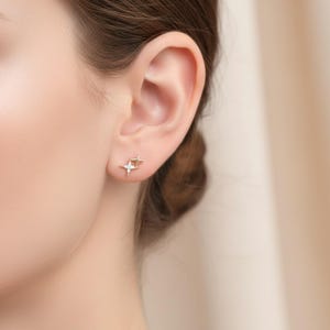 May include: A close-up of an ear adorned with two small, gold-coloured star-shaped stud earrings. One earring has a small, clear gemstone at its centre. The earrings are set against a blurred, neutral-toned background.