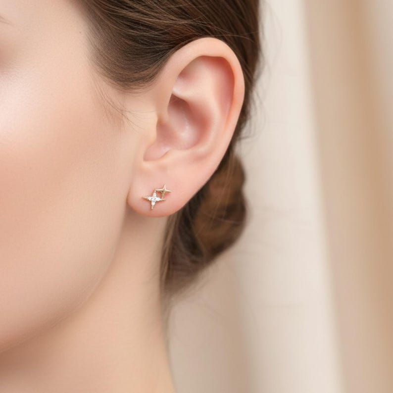 May include: A close-up of an ear adorned with two small, gold-coloured star-shaped stud earrings. One earring has a small, clear gemstone at its centre. The earrings are set against a blurred, neutral-toned background.
