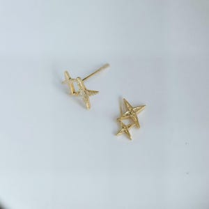May include: Gold-coloured star-shaped stud earrings. One earring features a single star with a small, clear gem in the centre. The other earring has two stars. The earrings are on a white background.