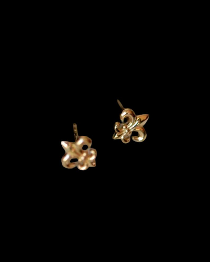 K&ouml;nnte beinhalten: Ein Paar goldfarbene Ohrstecker, jeweils in Form einer Fleur-de-Lis. Die Ohrringe sind vor einem schwarzen Hintergrund platziert, der ihr filigranes Design und ihren metallischen Glanz hervorhebt. Ein klassisches und elegantes Schmuckst&uuml;ck.