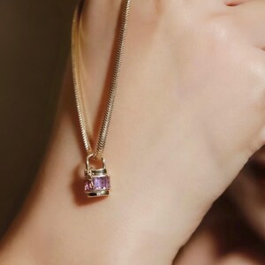 May include: A gold-colored necklace with a rectangular pendant featuring purple gemstones. The chain has a textured, snake-like design. The pendant is a small, decorative lock shape. The necklace is worn on a person's neck.