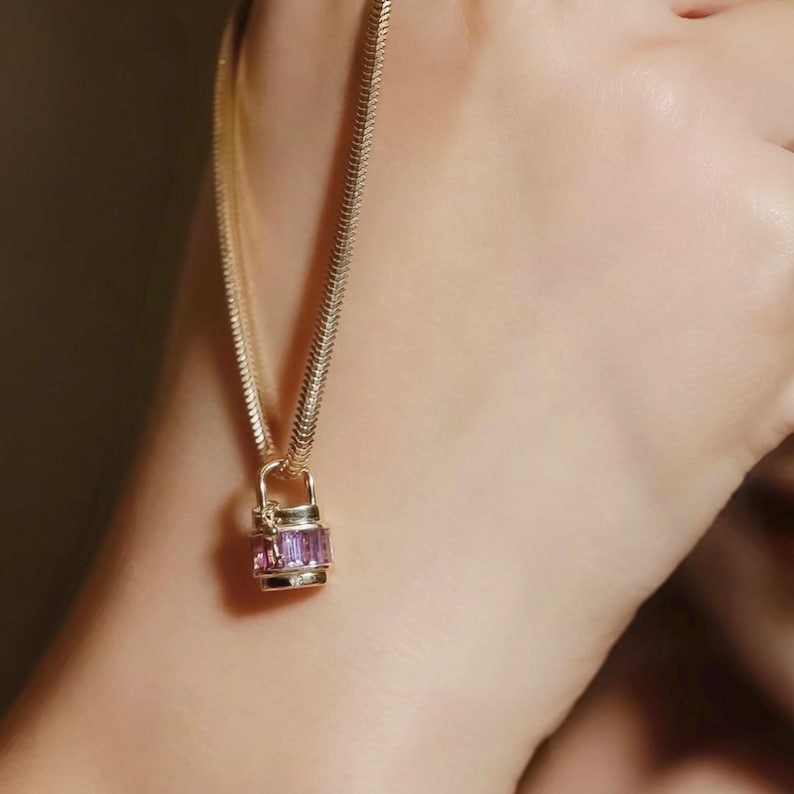 May include: A gold-colored necklace with a rectangular pendant featuring purple gemstones. The chain has a textured, snake-like design. The pendant is a small, decorative lock shape. The necklace is worn on a person's neck.