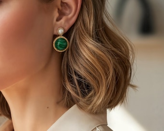 May include: Gold-toned earrings with a round, green stone and a small, white pearl. The green stone is surrounded by a gold-colored border. The earrings are a classic, elegant style.