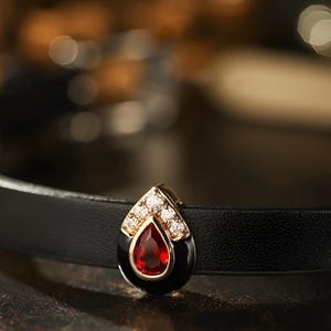Puede incluir: Un collar gargantilla de cuero negro con un colgante dorado que presenta una piedra roja rodeada de diamantes de imitación transparentes.