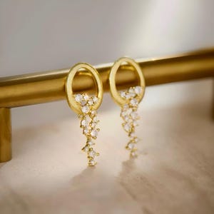 Peut inclure: Boucles d'oreilles créoles dorées avec une chaîne pendante de petites pierres claires.