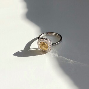 Puede incluir: Un anillo de plata con una piedra central de forma cuadrada. La piedra central es un diamante amarillo rodeado de diamantes blancos m&aacute;s peque&ntilde;os.