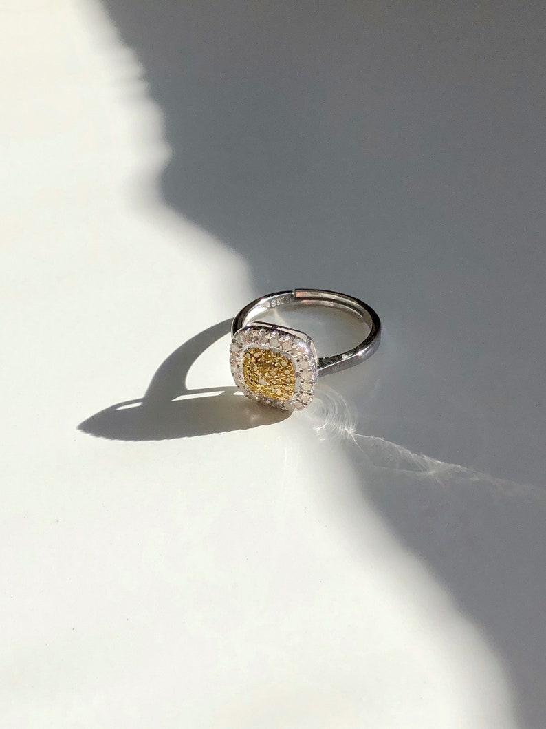 Puede incluir: Un anillo de plata con una piedra central de forma cuadrada. La piedra central es un diamante amarillo rodeado de diamantes blancos m&aacute;s peque&ntilde;os.