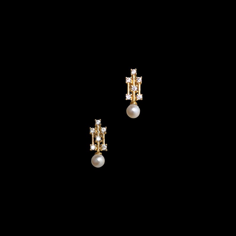 May include: A pair of gold earrings featuring a vertical rectangular frame set with four small clear stones, with a single round white pearl suspended from the base of each earring, presented against a solid black background.