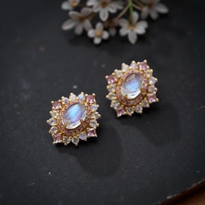 Puede incluir: Dos pendientes de bot&oacute;n dorados con centros ovalados de piedra lunar iridiscente. Los pendientes est&aacute;n rodeados de gemas rosas y blancas, creando un dise&ntilde;o floral. Sobre un fondo oscuro.