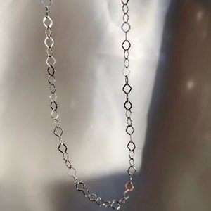 May include: A silver-toned necklace with a geometric diamond-shaped link design. The necklace is displayed against a soft, neutral background. A single gold-toned link adds a subtle contrast to the design.