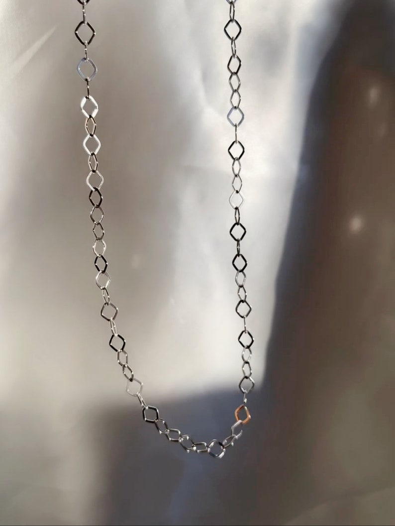 May include: A silver-toned necklace with a geometric diamond-shaped link design. The necklace is displayed against a soft, neutral background. A single gold-toned link adds a subtle contrast to the design.