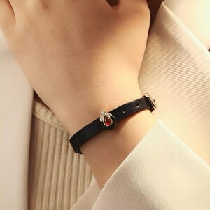 May include: A black leather bracelet with a gold charm featuring a red gemstone and clear rhinestones.